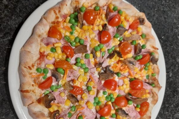 Pizza with ham, peas, sweetcorn, mushrooms and tomatoes on a white plate.